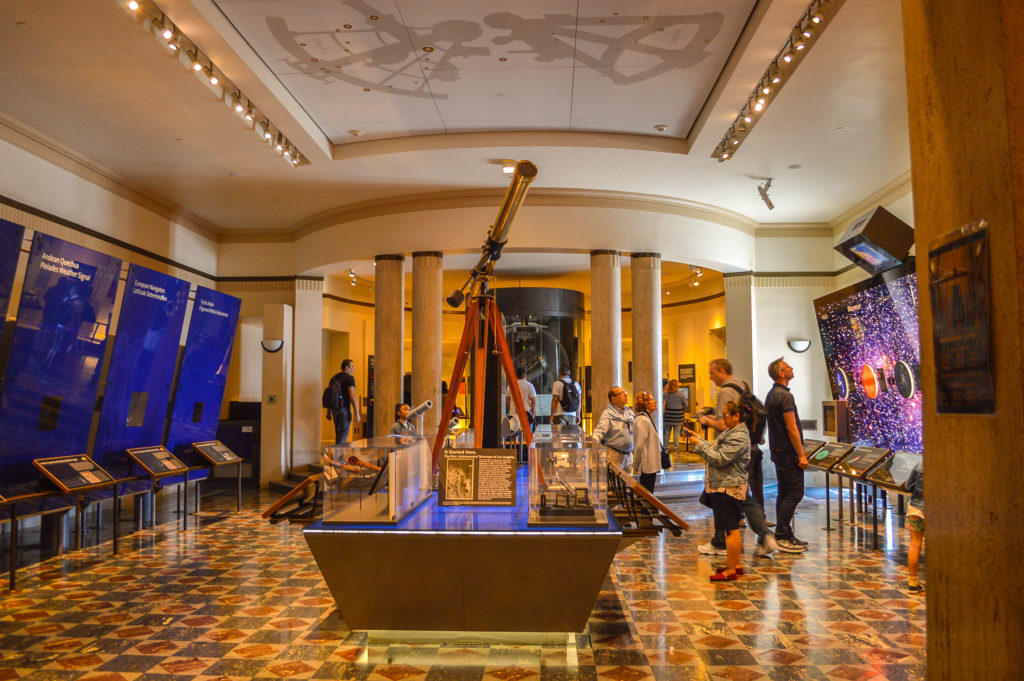 Interior of Griffith Observatory museum wide angle shot showing telescope on table in center with exhibits on the right and left of a rectangular room